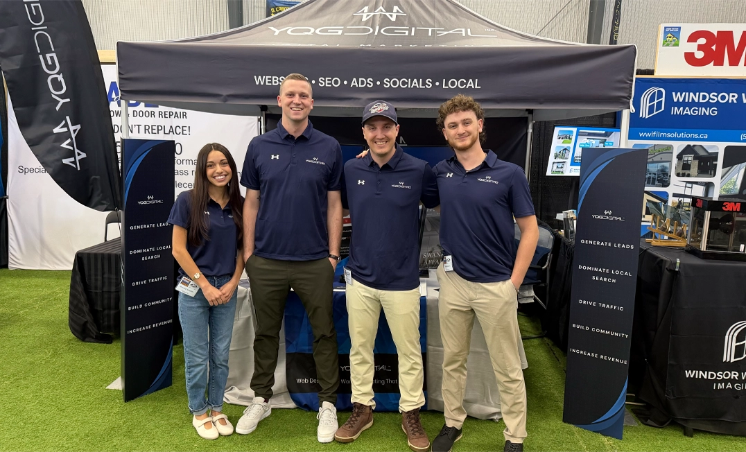 Four people in matching navy polo shirts pose for a photo at a trade show booth, standing on artificial turf with banners and displays behind them.