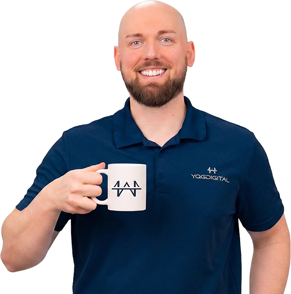 Smiling man with a shaved head and beard, wearing a navy polo, holds a white mug with a logo toward the camera.