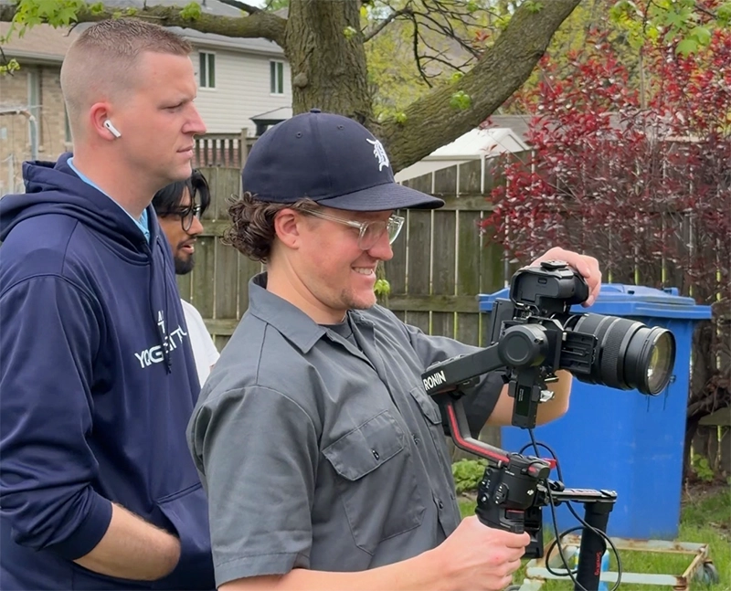 Cameraman adjusts a large video camera on a stabilizer outdoors while two people observe in a backyard setting with a blue recycling bin nearby.